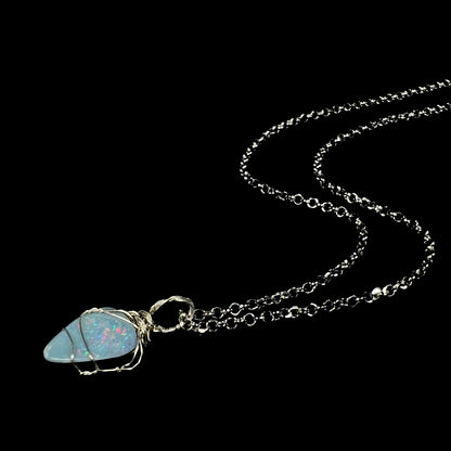 A women's freeform-shaped sterling silver wire wrapped opal doublet necklace shown from an angle laying on a black background, its chain flowing.
