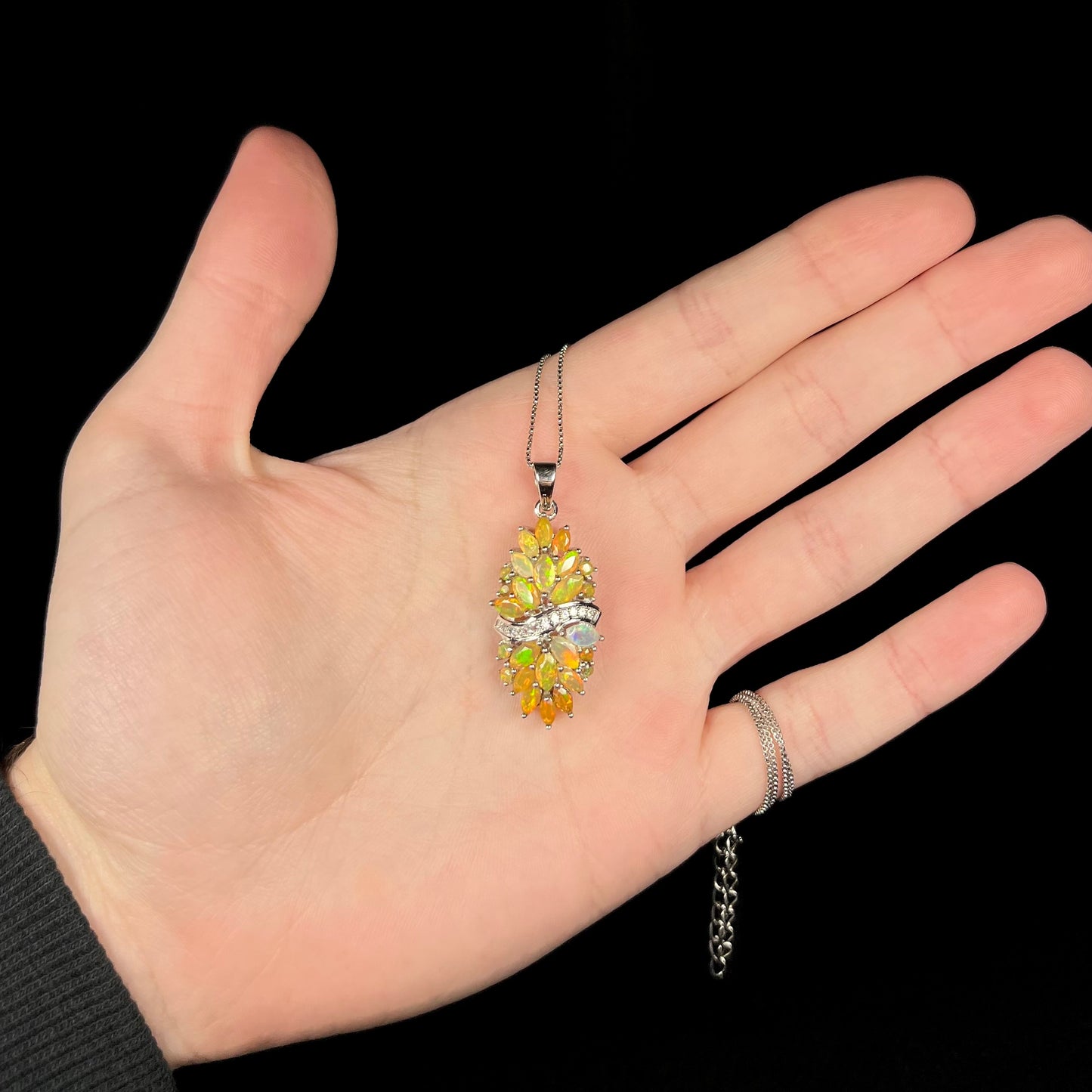 Hand holding a women's sterling silver Ethiopian fire opal cluster pendant against black background.  The necklace has a silver box chain wrapped around the pinky finger.