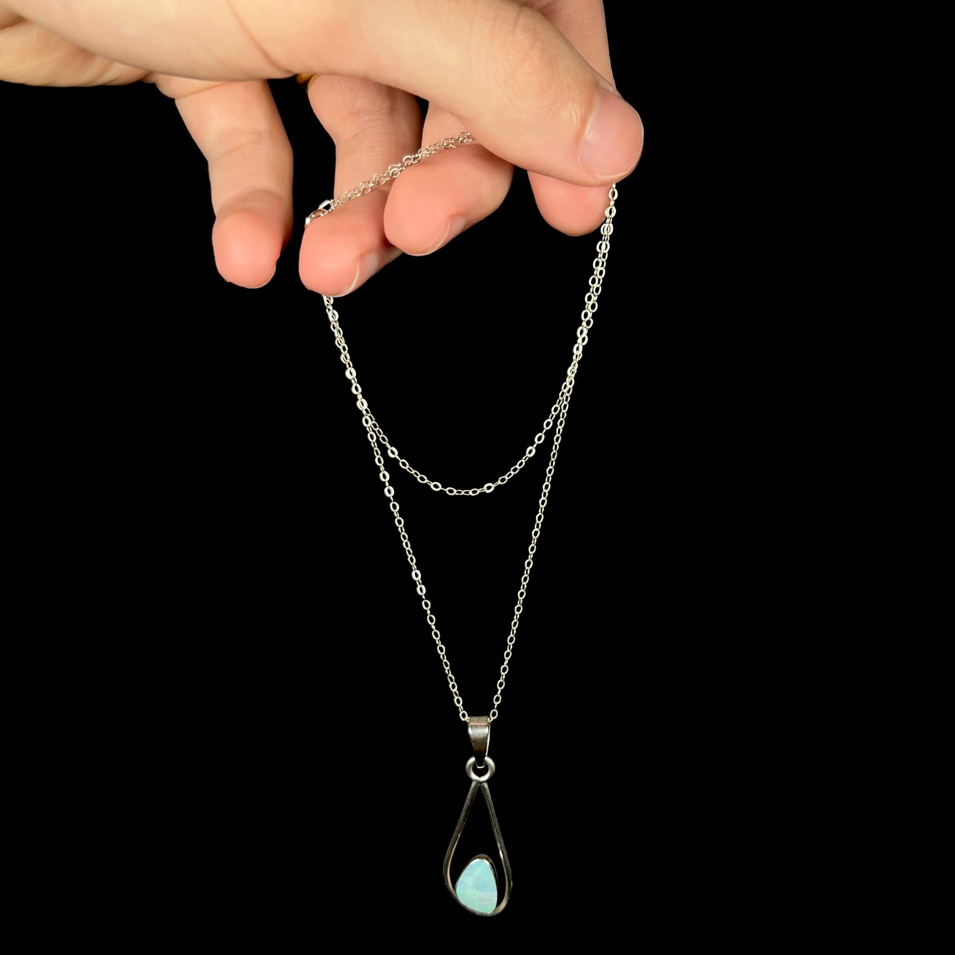 Hand dangling a women's sterling silver opal doublet necklace from its cable chain against a black background.
