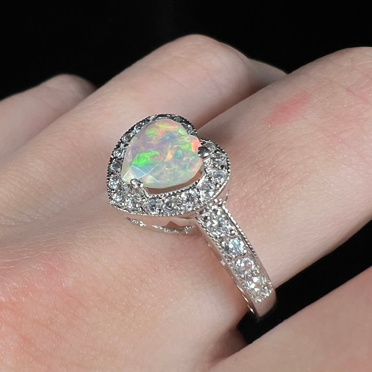 Angled view of women's sterling silver opal heart ring with milgrain details and pave set CZs, worn on finger