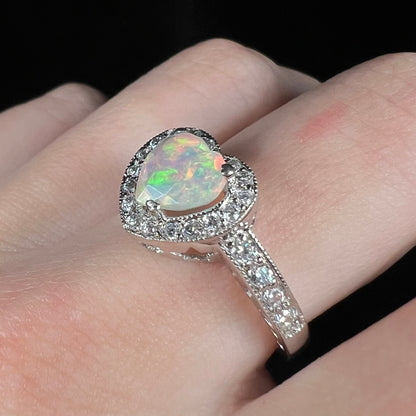 Angled view of women's sterling silver opal heart ring with milgrain details and pave set CZs, worn on finger