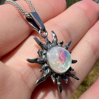 Close-up angled view of a women's sterling silver sun pendant set with a solitaire imitation opal, held in sunlight in a hand.  The opal has an iridescent hue.