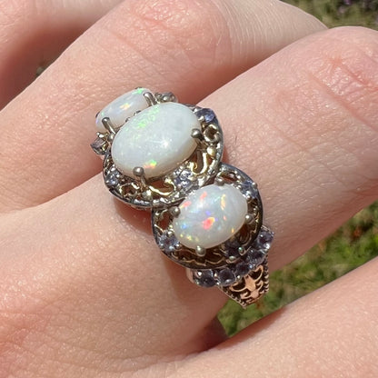 Angled close-up of a women's three-stone Australian opal and tanzanite-accented ring with filigree details, worn on a fingere in sunlight.  The opals have red and green colors.