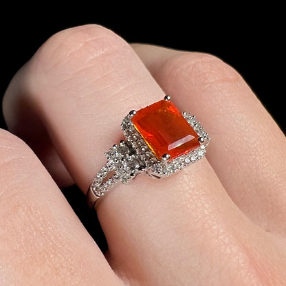 Women's white gold emerald cut fire opal and zircon ring shown close-up on a finger from an angle under cool studio lighting against a black background.