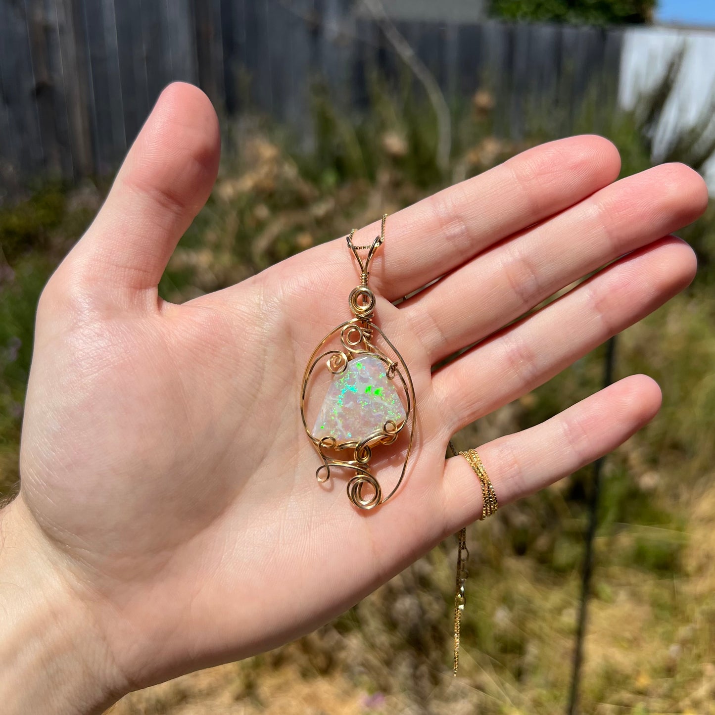Hand holding a women's gold-plated wire wrapped Australian crystal opal necklace in natural light against blurred outdoor backdrop.