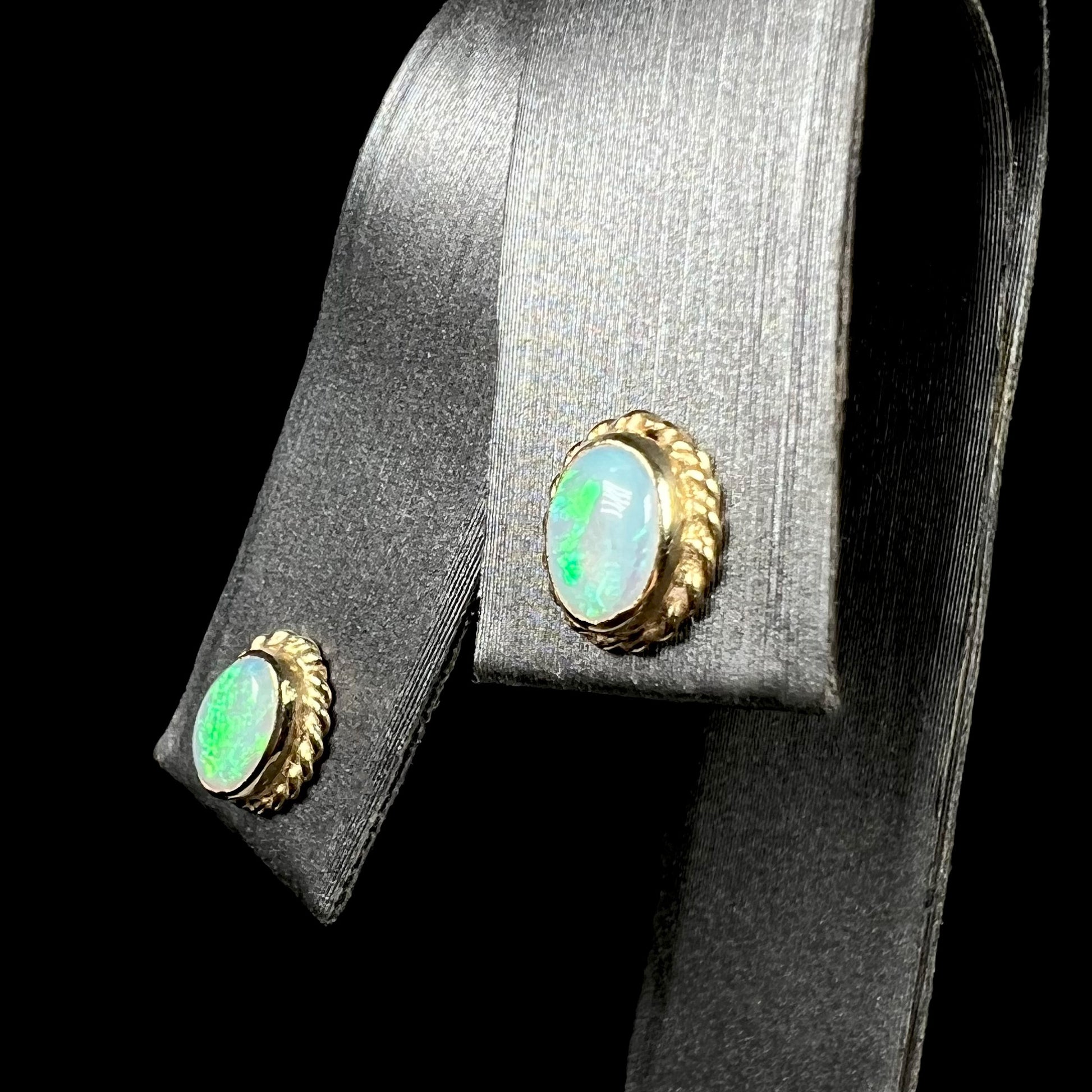 Pair of yellow gold opal stud earrings with green flashes shown from angle on earring display against black backdrop.