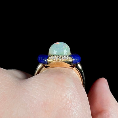 Side gallery profile view of a high-domed Ethiopian opal, diamond, and lapis lazuli ring in yellow gold, worn on a hand against a black background.