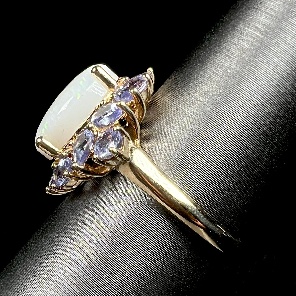 Side view of a yellow gold white opal and tanzanite-accented ring, shown on ring display; the ring's lightweight yellow gold shank is in focus