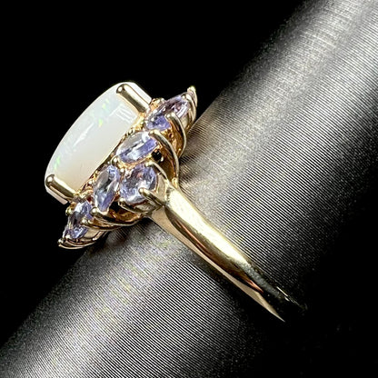 Side view of a yellow gold white opal and tanzanite-accented ring, shown on ring display; the ring's lightweight yellow gold shank is in focus