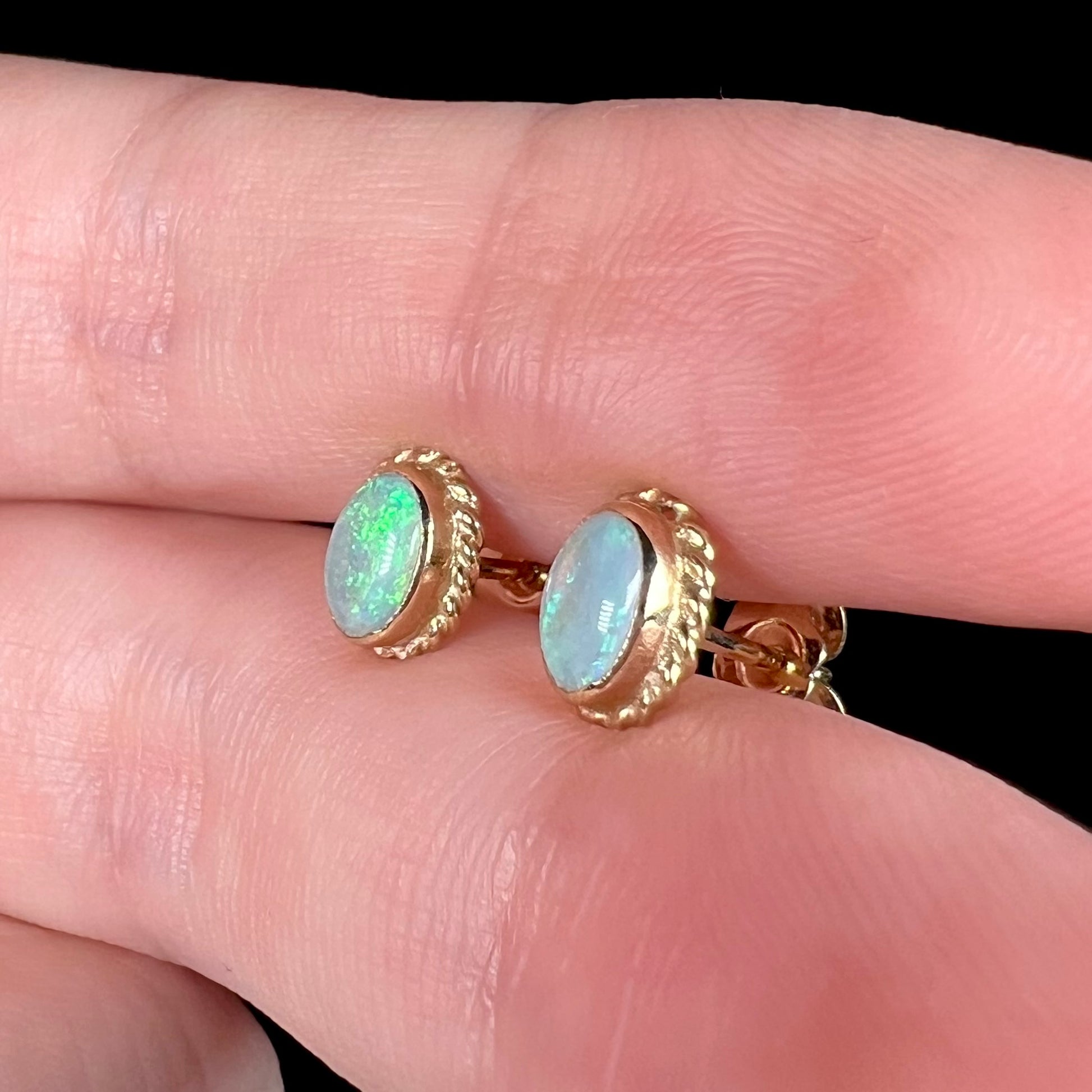 Pair of yellow gold rope design stud earrings set with opals shown close-up from the side held between two fingers.