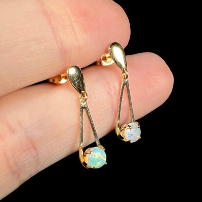 Angled view of round opal drop earrings held between two fingers against black background.