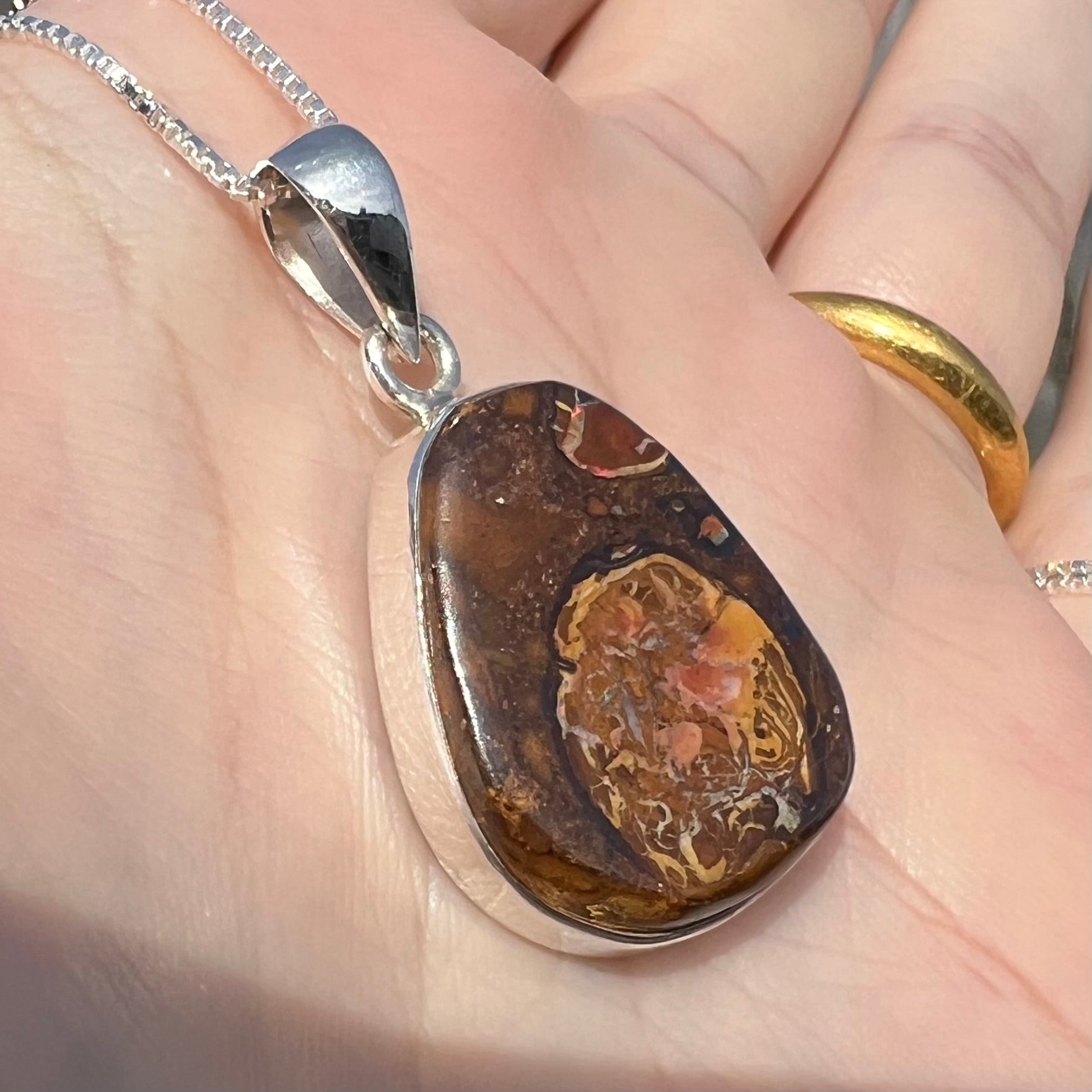 Angled close-up of a Yowah nut boulder opal pendant in sterling silver, shown in the sunlight held in a hand.